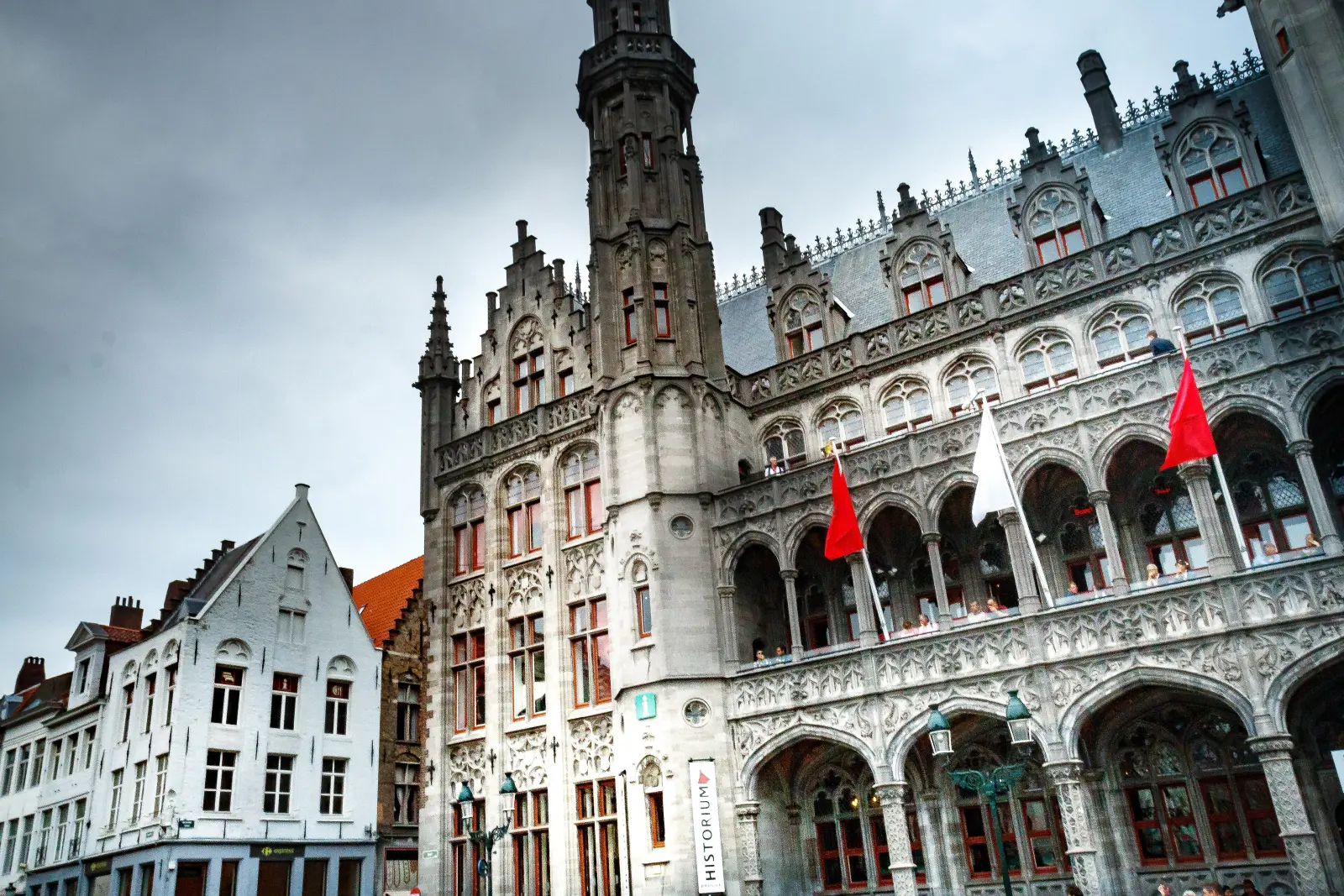 L'Historium sul Markt a Bruges: un edificio neogotico grigio con una slanciata torre angolare e bandiere rosse sulla facciata ad arcate